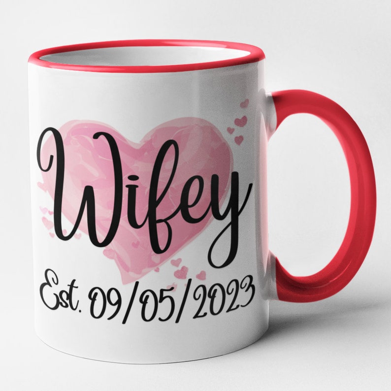 May include: White ceramic mug with a red rim and handle. The mug features a pink watercolor heart with the word "Wifey" in black script. The date "Est. 09/05/2023" is written below the heart.