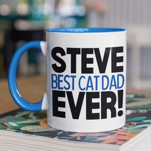 May include: White ceramic mug with a blue rim and handle. The mug has black text that reads "STEVE BEST CAT DAD EVER!"