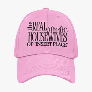 May include: A pink baseball cap with a curved peak. The cap displays the text "the REAL HOUSEWIVES of 'INSERT PLACE'" in black, with a cityscape graphic above the word "HOUSEWIVES".