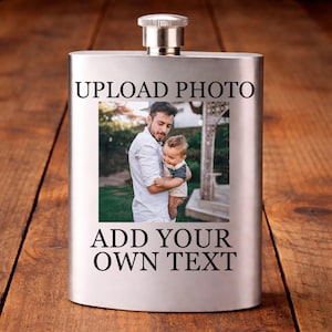 May include: A silver hip flask with a shiny metal cap. The flask is rectangular and displays a photo of a man holding a baby. The text "UPLOAD PHOTO" and "ADD YOUR OWN TEXT" are printed on the flask.