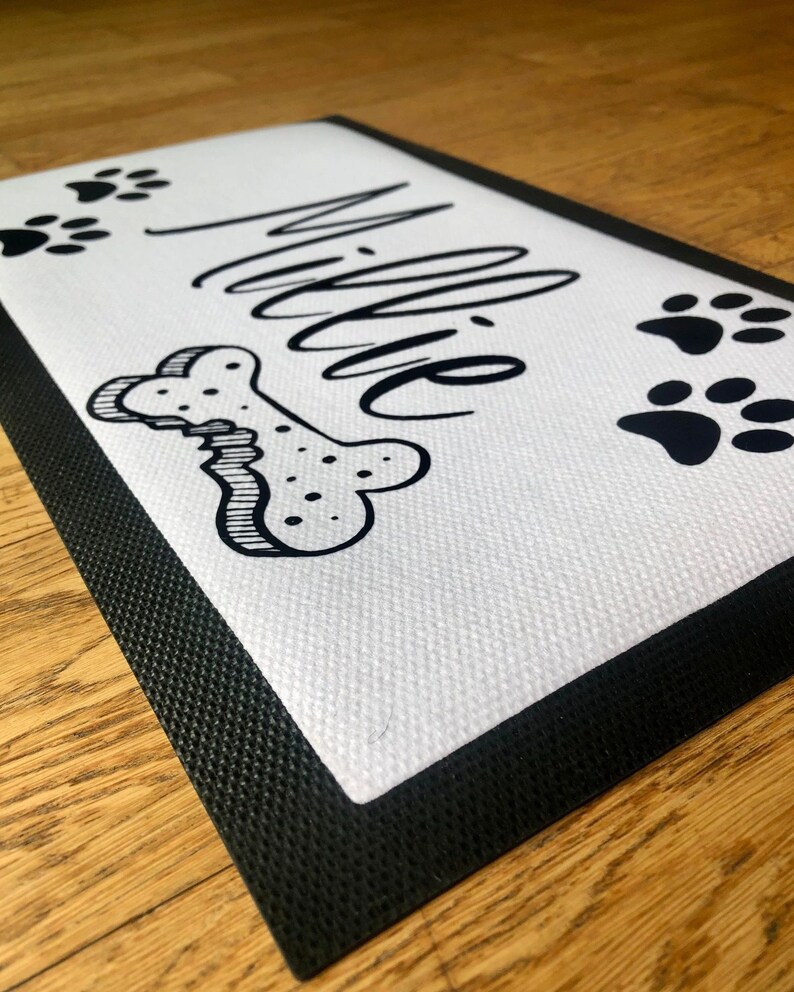 May include: White doormat with black border and black paw prints and bone design. The name "Millie" is written in black cursive.