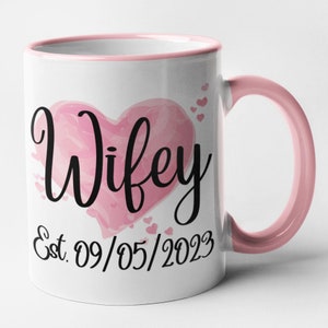 May include: White ceramic mug with a pink rim and handle. The mug features a pink watercolor heart with the word "Wifey" in black script. The date "Est. 09/05/2023" is written below the heart.