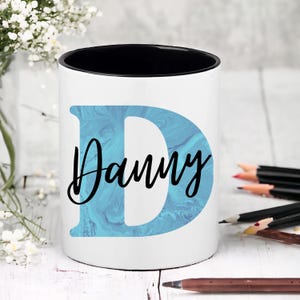 May include: White ceramic mug with a black rim. The mug features a blue and white marbled letter "D" and the name "Danny" written in black cursive.