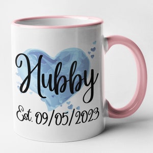 May include: White ceramic mug with a pink rim and handle. The mug features a blue watercolor heart with the word "Hubby" in black script. The date "Est. 09/05/2023" is printed below the heart.
