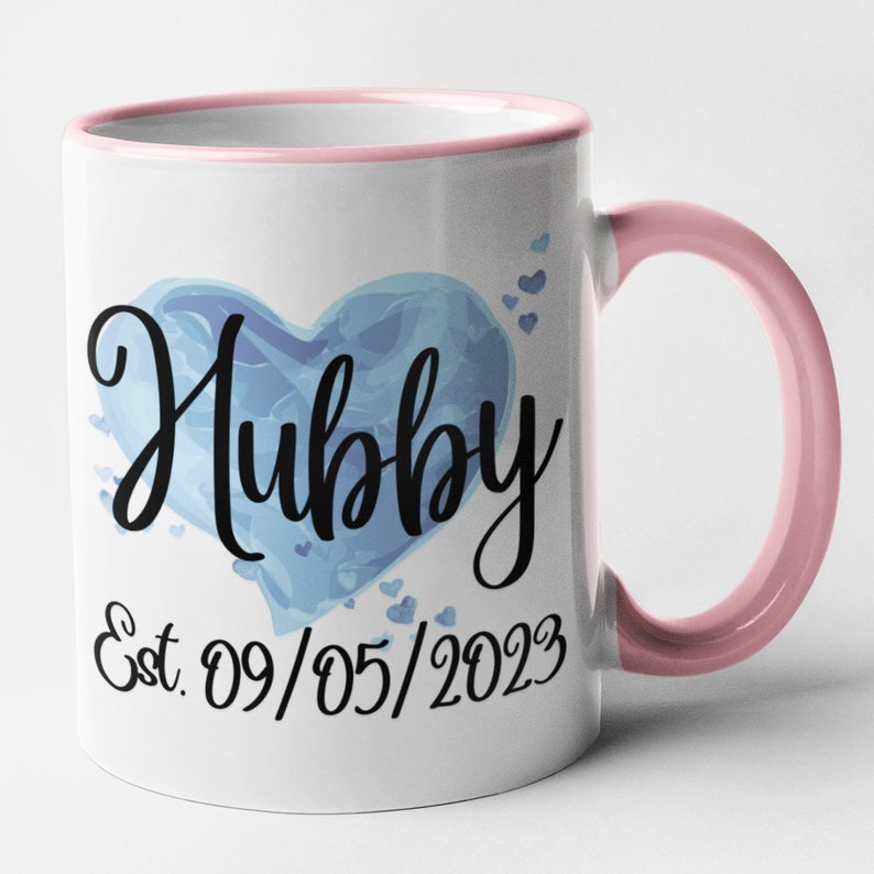 May include: White ceramic mug with a pink rim and handle. The mug features a blue watercolor heart with the word "Hubby" in black script. The date "Est. 09/05/2023" is printed below the heart.