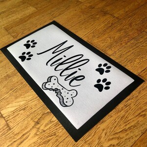 May include: A white doormat with a black border and the name "Millie" written in cursive. There are three black paw prints and a black bone with a bite mark on the doormat.