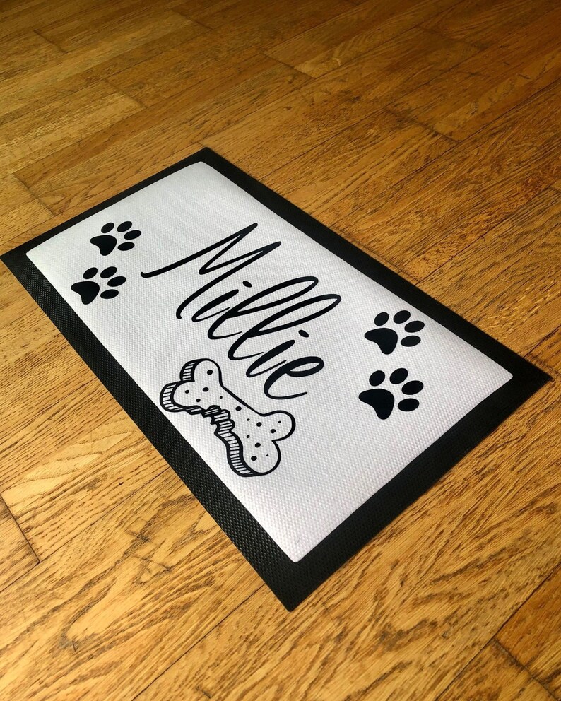 May include: A white doormat with a black border and the name "Millie" written in cursive. There are three black paw prints and a black bone with a bite mark on the doormat.
