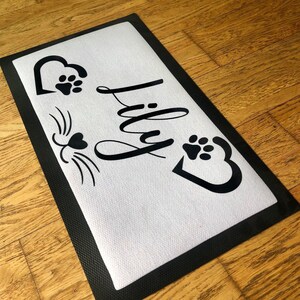 May include: Black and white doormat with the name "Lily" written in cursive. The doormat has a black border and a white background. There are two paw prints and a heart on the doormat.