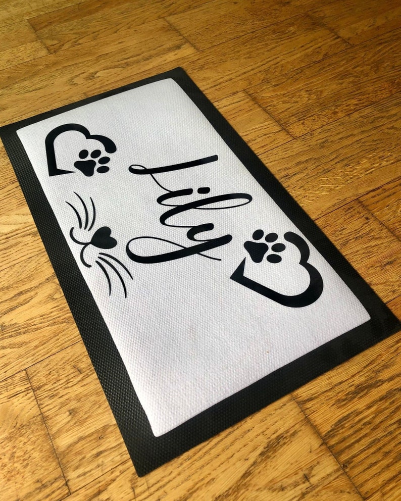 May include: Black and white doormat with the name "Lily" written in cursive. The doormat has a black border and a white background. There are two paw prints and a heart on the doormat.