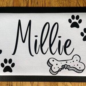 May include: White doormat with black border and black paw prints, a bone, and the name "Millie" written in cursive.