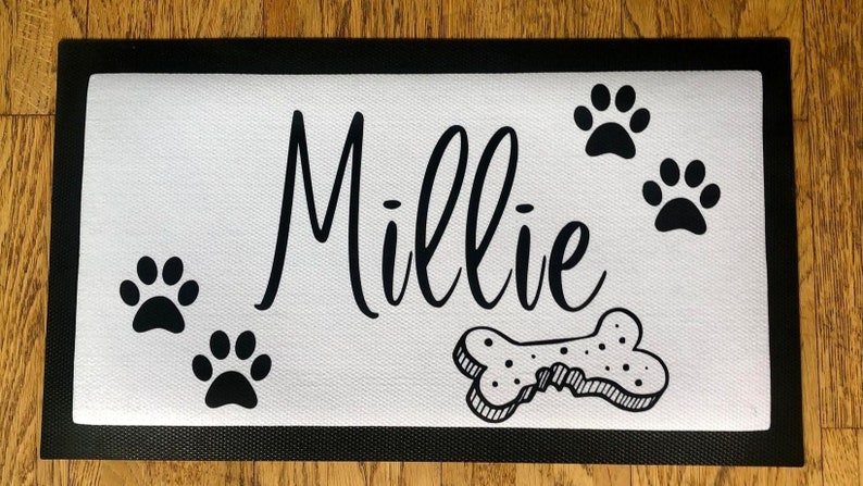 May include: White doormat with black border and black paw prints, a bone, and the name "Millie" written in cursive.