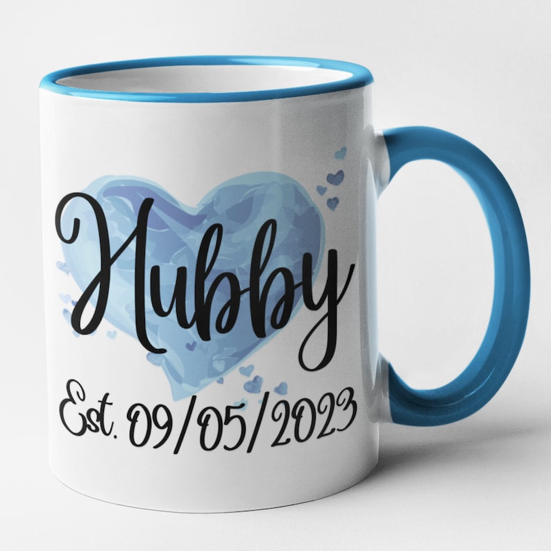 May include: White ceramic mug with a blue rim and handle. The mug features a blue watercolor heart with the word "Hubby" in black script. The date "Est. 09/05/2023" is printed below the heart.