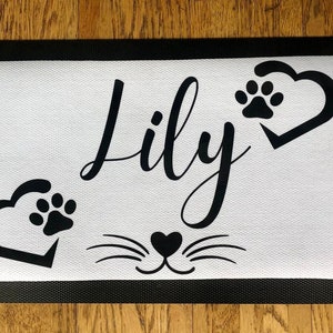 May include: Black and white doormat with the name "Lily" in cursive script. The doormat has two paw prints inside hearts and a cat face with whiskers.