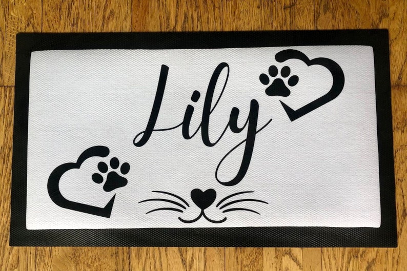 May include: Black and white doormat with the name "Lily" in cursive script. The doormat has two paw prints inside hearts and a cat face with whiskers.