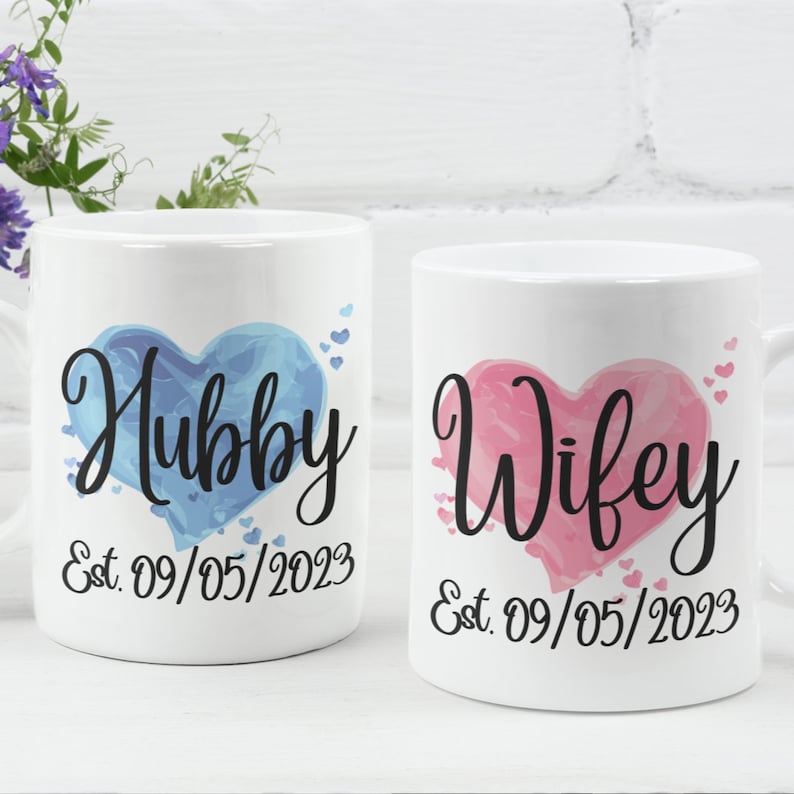May include: Two white ceramic mugs with blue and pink watercolor hearts. The mug on the left says "Hubby Est. 09/05/2023" and the mug on the right says "Wifey Est. 09/05/2023".