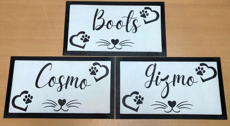 May include: Three black framed white mats with black paw print hearts and a cat face design. The mats have the names "Boots", "Cosmo", and "Gizmo" written in black cursive.