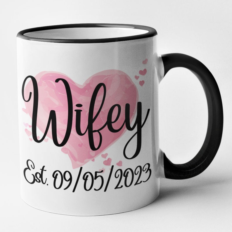 May include: White ceramic mug with a black rim and handle. The mug features a pink watercolor heart with the word "Wifey" in black script. Below the heart is the text "Est. 09/05/2023".