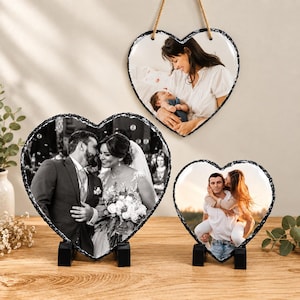 May include: Three heart-shaped slate plaques with rough, dark edges. One plaque hangs by a rope, displaying a person holding a baby. Two plaques stand on black bases, one showing a wedding couple and the other a person giving a piggyback ride.