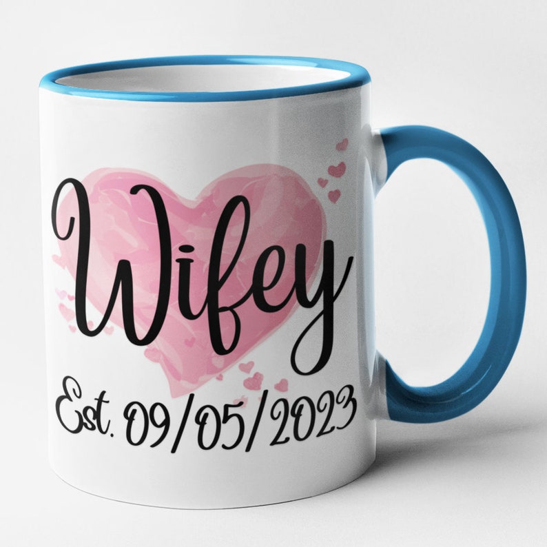 May include: White ceramic mug with a blue rim and handle. The mug features a pink watercolor heart with the word "Wifey" in black script. Below the heart is the text "Est. 09/05/2023" in black.