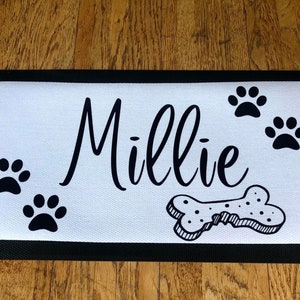 May include: White doormat with black border and black paw prints, a bone, and the name "Millie" written in cursive.