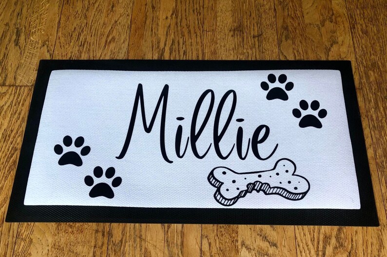 May include: White doormat with black border and black paw prints, a bone, and the name "Millie" written in cursive.