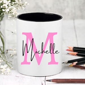 May include: White ceramic pencil holder with a black rim. The holder features a large pink letter "M" and the word "Michelle" written in black cursive script.