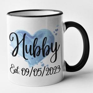 May include: White ceramic mug with a black rim and handle. The mug features a blue watercolor heart with the word "Hubby" in black script. Below the heart is the text "Est. 09/05/2023".