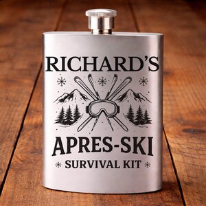 May include: A stainless steel flask with a silver cap. The flask is engraved with the text "RICHARD'S APRES-SKI SURVIVAL KIT" along with a graphic of skis, goggles, mountains, and trees. The flask is rectangular and sits on a wooden surface.