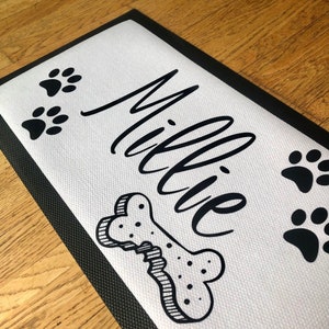 May include: Black and white dog doormat with the name "Millie" written in cursive, a bone, and paw prints.