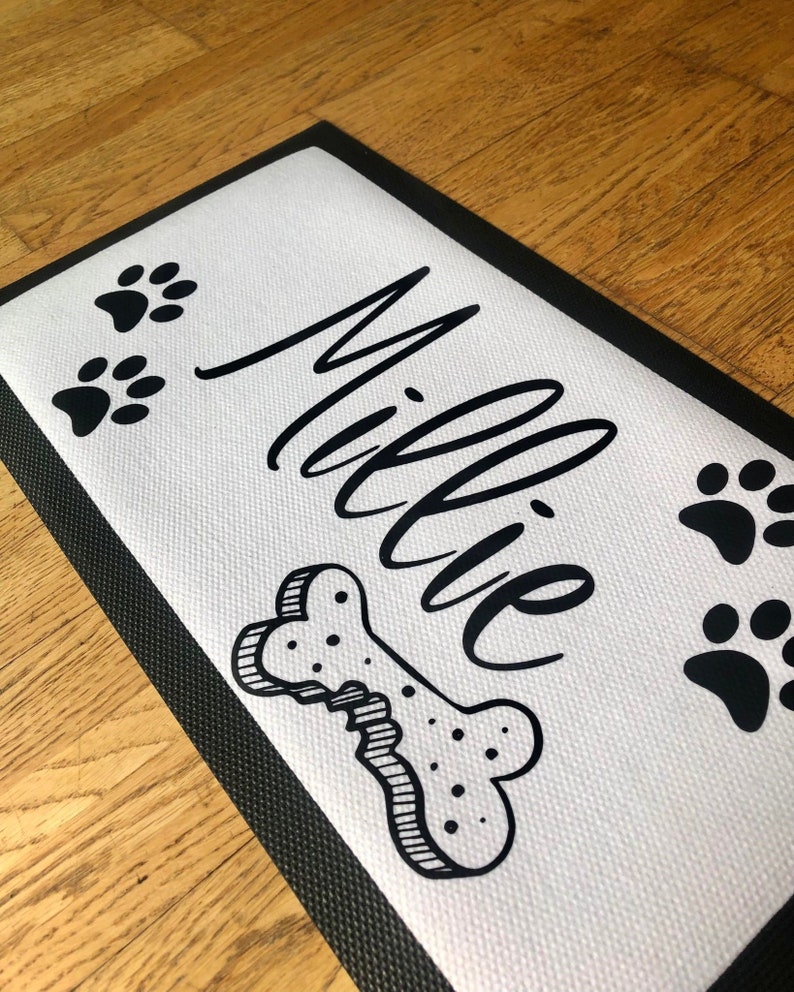 May include: Black and white dog doormat with the name "Millie" written in cursive, a bone, and paw prints.