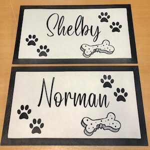 May include: Two black and white dog doormats with black borders. The top mat says "Shelby" and the bottom mat says "Norman". Both mats have a bone and paw prints.