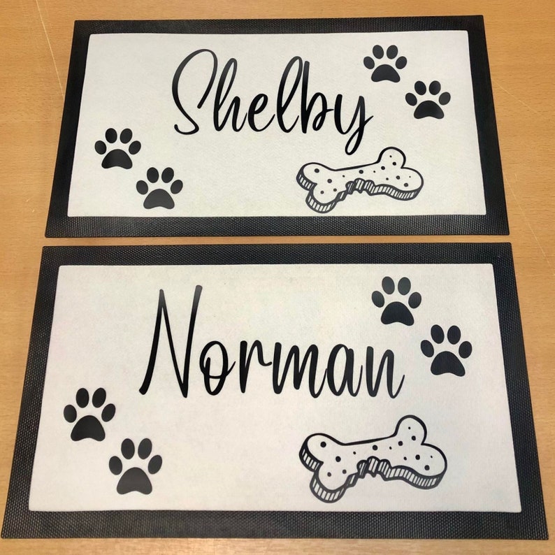 May include: Two black and white dog doormats with black borders. The top mat says "Shelby" and the bottom mat says "Norman". Both mats have a bone and paw prints.