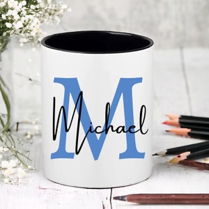 May include: White ceramic pencil holder with a black rim. The holder features a blue letter "M" and the name "Michael" in black script.