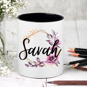 May include: White ceramic pencil holder with a black rim. The holder features a gold geometric frame with purple and pink floral accents and the name "Savannah" in black script.