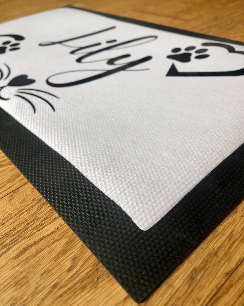 May include: White doormat with a black border and the name "Lily" written in a cursive font.  Two paw prints and a cat face are also printed on the mat.
