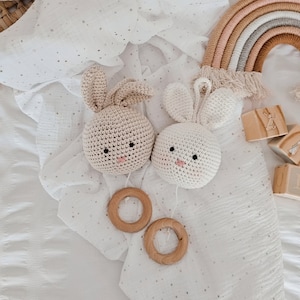 Op de afbeelding: Twee gehaakte konijnenrammelaars met houten ringen die hangen aan een wit muslin deken met kleine witte stippen. De konijnen zijn beige en wit met zwarte ogen en roze neuzen. Een regenboogkleurige macramé wandhanger is op de achtergrond.
