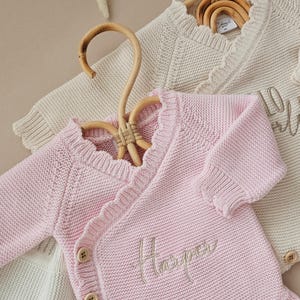 May include: Two long-sleeved knit bodysuits with a wrap-style front and ruffled trim along the neckline. The pink bodysuit features the embroidered name Harper and three wooden buttons on the side. The cream bodysuit has a similar design.
