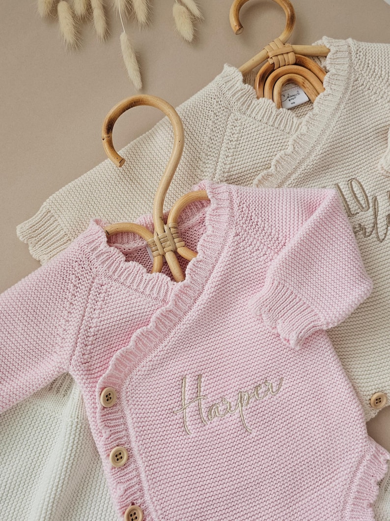 May include: Two long-sleeved knit bodysuits with a wrap-style front and ruffled trim along the neckline. The pink bodysuit features the embroidered name Harper and three wooden buttons on the side. The cream bodysuit has a similar design.
