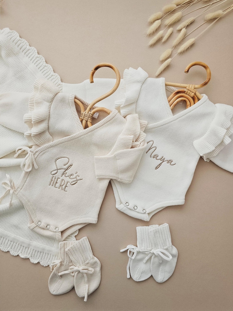 Personalised Baby Ruffle Romper in white, made from 100% cotton. Luxury neutral design with optional matching headband and booties. Custom embroidery options: 'Hello World,' 'She’s Here,' 'Isn’t She Lovely,' or baby’s name. Perfect for newborn coming home outfits, birth announcements, and baby shower gifts. Available in newborn size (fits up to 2-3 months). Matching rattle sold separately. Hand wash only. Ideal for baby girl outfits. Customizable baby romper set.