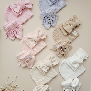 May include: A collection of knitted baby sets in various pastel colours, each including a beanie with a large bow, a headband with a bow, and a pair of booties. The sets are arranged in shades of pink, light blue, beige, cream, and white, all featuring a small white label.