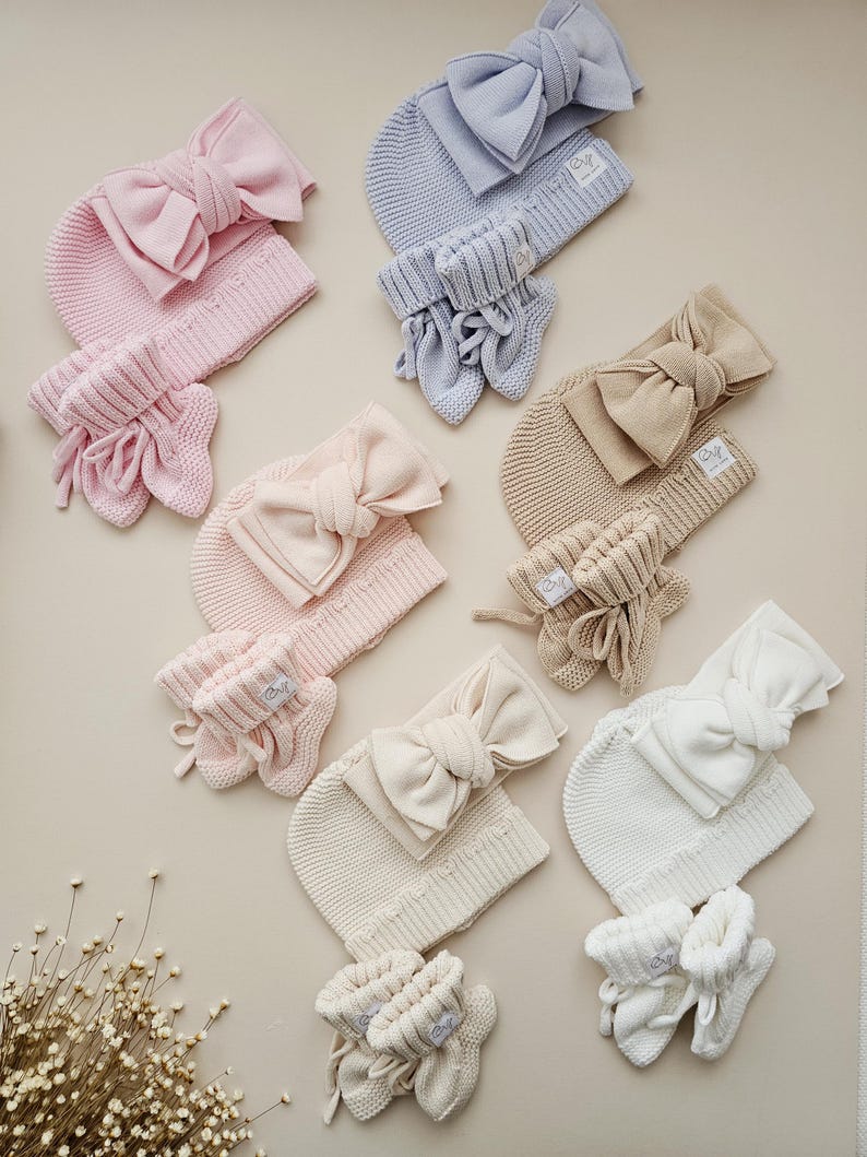 May include: A collection of knitted baby sets in various pastel colours, each including a beanie with a large bow, a headband with a bow, and a pair of booties. The sets are arranged in shades of pink, light blue, beige, cream, and white, all featuring a small white label.