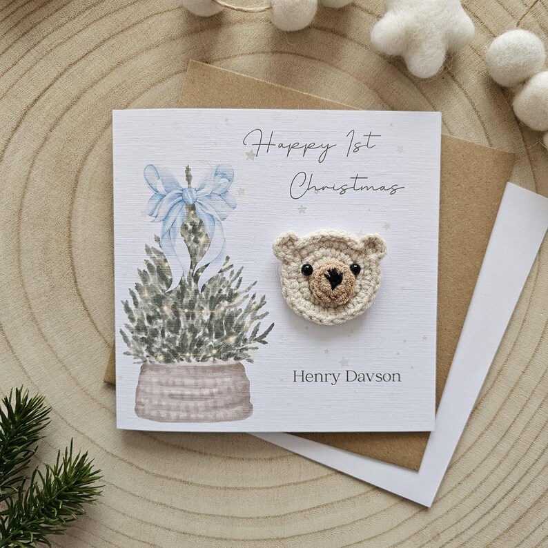 May include: A Christmas card featuring a watercolour illustration of a Christmas tree and a crocheted teddy bear head. The card reads "Happy 1st Christmas" and includes a name. The card is on a wooden surface.