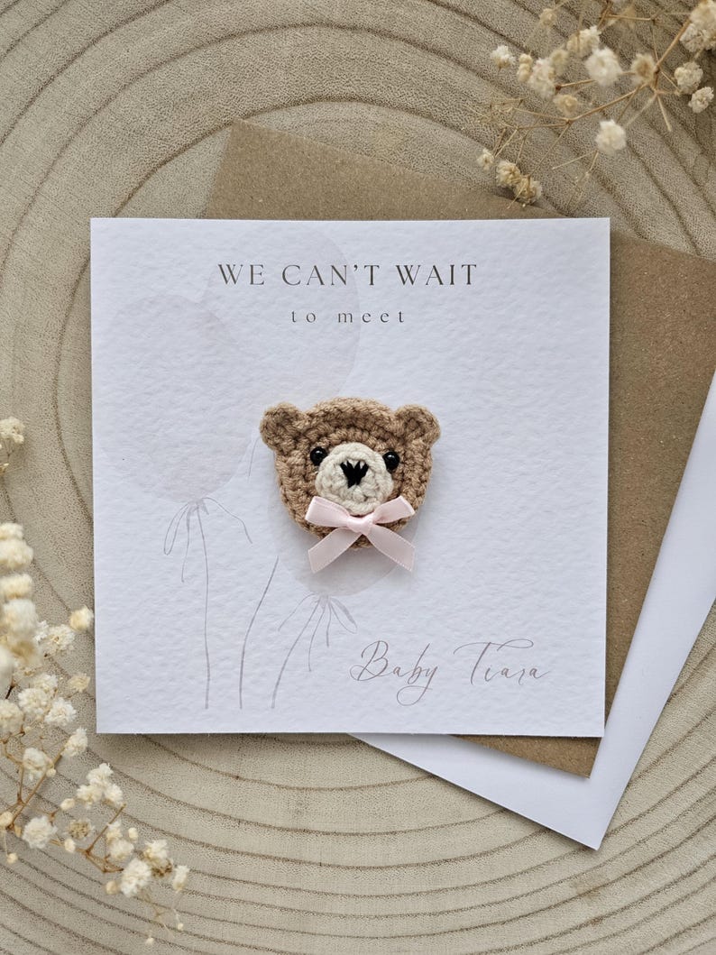 May include: A handmade baby announcement card with a crocheted teddy bear head and a pink bow. The card reads "WE CAN'T WAIT to meet Baby Tiara" in elegant script. The card is on a wooden surface.