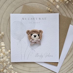 May include: A greeting card with a crocheted teddy bear attached. The card reads "WE CAN'T WAIT to meet" and "Baby John". The bear is tan with a cream-colored bow. The card is on a wooden surface.