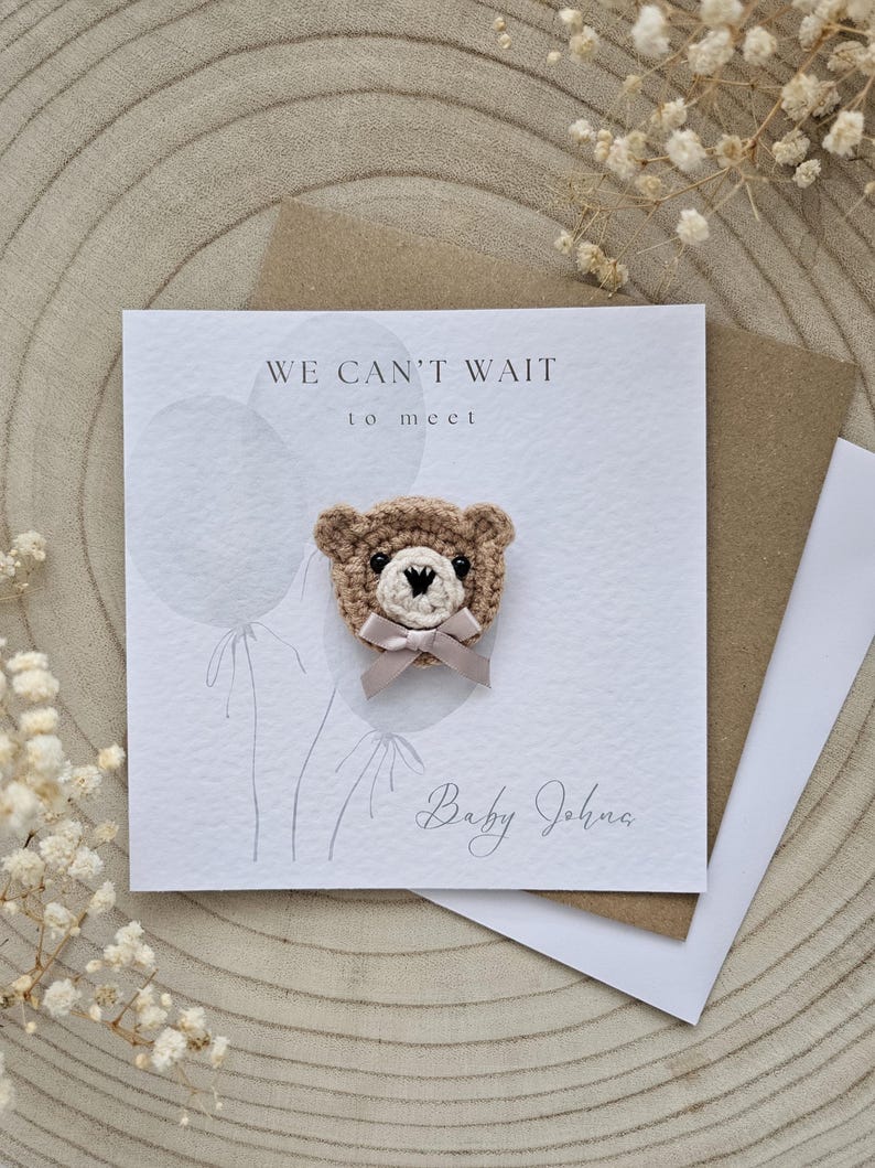 May include: A greeting card with a crocheted teddy bear attached. The card reads "WE CAN'T WAIT to meet" and "Baby John". The bear is tan with a cream-colored bow. The card is on a wooden surface.