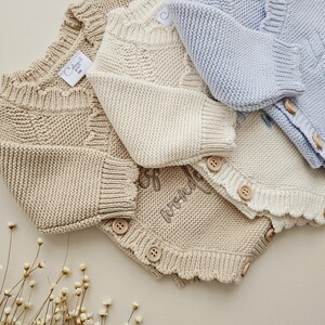 May include: Three knitted baby rompers in light blue, cream, and tan. Each romper features a ribbed neckline, long sleeves, and wooden buttons along the front and leg openings. The cream romper displays the embroidered text "full of wonder".