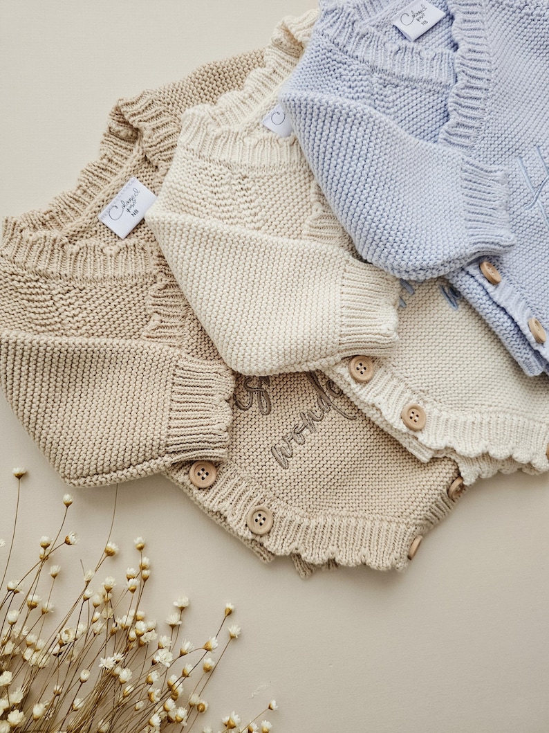 May include: Three knitted baby rompers in light blue, cream, and tan. Each romper features a ribbed neckline, long sleeves, and wooden buttons along the front and leg openings. The cream romper displays the embroidered text "full of wonder".