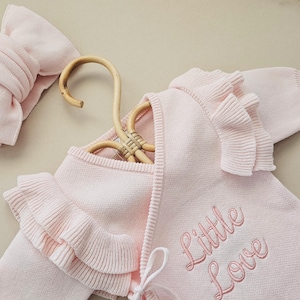 May include: A light pink baby sweater with ruffled edges and the embroidered text "Little Love". A matching headband is visible on the left. The sweater is displayed on a wooden hanger.