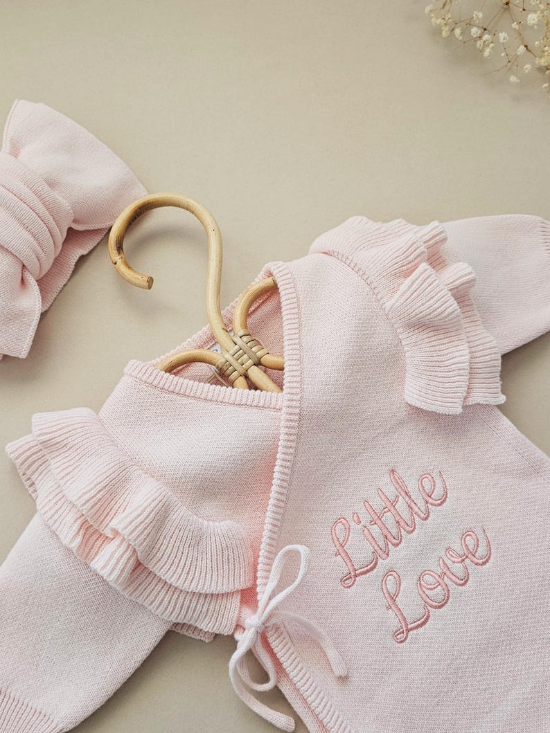 May include: A light pink baby sweater with ruffled edges and the embroidered text "Little Love". A matching headband is visible on the left. The sweater is displayed on a wooden hanger.