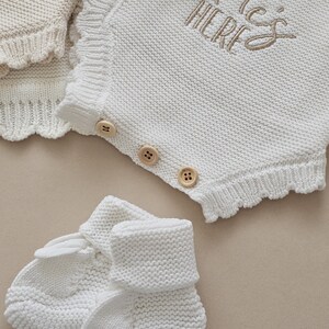 Puede incluir: Un conjunto de ropa de beb&eacute; de punto blanco, que incluye un pelele con botones de madera y el texto bordado "He's HERE", y patucos a juego. El pelele tiene un borde festoneado. El conjunto est&aacute; hecho de tejido de punto suave y texturizado.